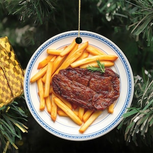 May include: A round ceramic ornament featuring a printed image of a seared steak and golden french fries on a white plate with a blue patterned rim. The ornament is suspended by a thin gold string and displays a realistic food photograph.