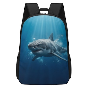 Puede incluir: Una mochila negra que presenta un gran estampado gráfico de alta resolución de un gran tiburón blanco nadando bajo el agua. El tiburón está rodeado de burbujas de aire sobre un fondo de océano azul profundo con rayos de luz filtrándose desde arriba.