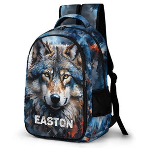 May include: A backpack featuring a watercolor-style illustration of a wolf's face with amber eyes. The design incorporates shades of blue, black, and orange. The name EASTON is printed in white capital letters at the bottom of the front pocket.