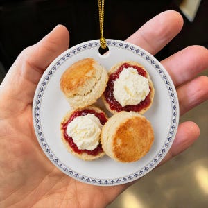 May include: A round ceramic ornament featuring a printed image of four golden-brown scones on a white plate. Two scones are topped with red jam and white clotted cream. The ornament has a blue and white decorative border and a gold hanging cord.