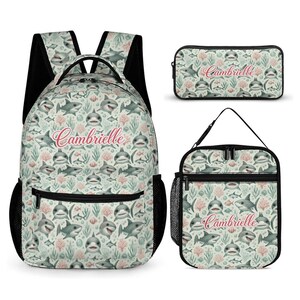 May include: A backpack, a rectangular pencil case, and a lunch bag featuring a repeating pattern of grey sharks, green seaweed, and pink coral on a light background. Each item displays the name Cambrielle in a pink cursive script font.