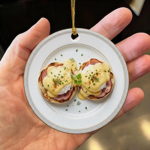 以下が含まれることがあります： ハムをのせたトースト済みのイングリッシュマフィンに、オランデーズソース、新鮮なチャイブ、ディルをトッピングしたエッグベネディクトが描かれた円形のセラミック製オーナメント。ゴールドカラーの吊り下げ用コードと装飾的な縁取りが施されています。