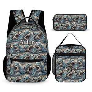 Puede incluir: Una mochila, un estuche rectangular y una bolsa para el almuerzo, todos con un patrón repetitivo de tiburones en aguas azules turbulentas. Cada artículo está personalizado con el nombre Aiden impreso en blanco en el centro del diseño.