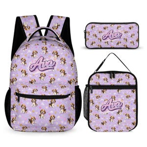 Op de afbeelding: Een set schoolspullen met een rugzak, etui en lunchtas. Ze hebben een lavendelkleurige achtergrond met een patroon van beaglepuppy's, wolken en harten. De naam "Ava" staat op elk item.