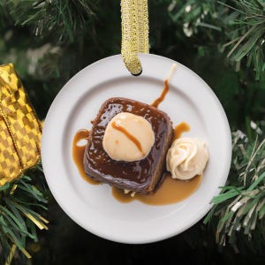Pode incluir: Um enfeite redondo de cerâmica branca com a imagem impressa de um pudim de caramelo quadrado num prato, coberto com uma porção de natas e molho de caramelo, suspenso por uma fita dourada para pendurar na árvore de Natal.