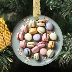 May include: A round, clear glass ornament featuring an assortment of colorful macarons. The macarons are in shades of pink, yellow, white, and purple, arranged on a light green surface with small pink floral accents. A gold ribbon is attached for hanging.