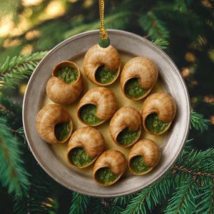 May include: A round, flat, beige ceramic ornament featuring a graphic of ten snail shells filled with green herb butter arranged in a circle on a plate. The ornament is suspended by a gold cord with a small green bead at the top.