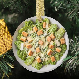 Puede incluir: Un adorno navideño redondo y plano que presenta una imagen de alta resolución de una ensalada César con lechuga romana verde crujiente, picatostes dorados y una ligera capa de queso parmesano rallado, suspendido por una cinta dorada.