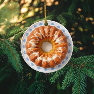 May include: A round, ceramic ornament shaped like a bundt cake with white icing. The ornament is on a white plate with a blue floral pattern. A gold string is attached for hanging, set against a backdrop of green pine needles.
