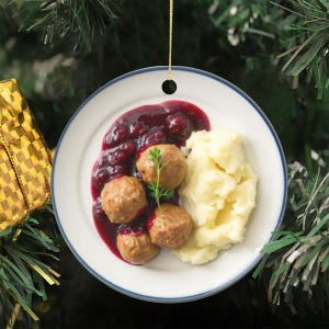 Puede incluir: Adorno navideño redondo con un plato de albóndigas suecas con salsa y puré de patatas. El plato tiene un borde azul y cuelga de un hilo dorado. El fondo es un árbol de Navidad.