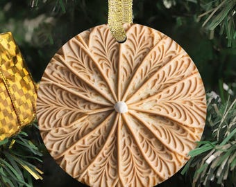Op de afbeelding: Een cirkelvormig beige ornament met een radiaal patroon van in reliëf aangebrachte varenachtige bladeren die vanuit een wit middelpunt naar buiten lopen. Het ornament hangt aan een glinsterend gouden lint en heeft een symmetrisch ontwerp.