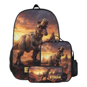 May include: A backpack, lunch bag, and pencil case set featuring a printed graphic of a roaring Tyrannosaurus Rex in a volcanic landscape at sunset. The name ETHAN is printed in gold block letters on the front of the pencil case and backpack.