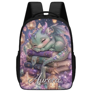 May include: A black backpack with a fantasy illustration of a sleeping dragon. The dragon is light green and rests on a stack of purple and gold books. The name "Aurora" is written in cursive at the bottom of the backpack.