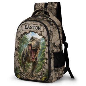 Puede incluir: Una mochila con un patrón de textura de piedra agrietada y un gráfico central de un Tyrannosaurus Rex rugiendo que emerge de una escena de selva. El nombre EASTON está impreso en letras negras de bloque sobre una pancarta de piedra arriba.