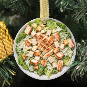 May include: Circular Christmas tree decoration with a Caesar salad design. The ornament depicts a white bowl filled with lettuce, croutons, and grilled chicken. A gold ribbon is attached for hanging on a Christmas tree.