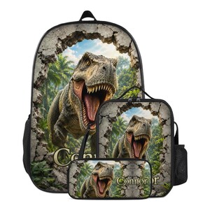 Pode incluir: Conjunto de mochila, lancheira e estojo pretos com um design de Tyrannosaurus Rex. O dinossauro parece estar quebrando uma parede de pedra. O nome "Connor" está impresso na lancheira e no estojo.