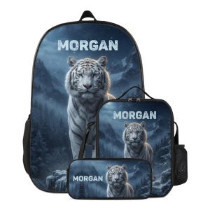 May include: A backpack, lunch bag, and pencil case set featuring a white tiger in a snowy mountain forest landscape. Each item displays the name MORGAN in white capital letters at the top. The set has black trim and mesh side pockets.