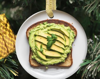May include: A round, white ceramic Christmas ornament featuring a realistic image of a slice of toasted bread topped with mashed avocado, sliced avocado pieces, and a small sprig of fresh green basil, suspended by a gold glitter ribbon.