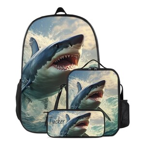 Puede incluir: Un conjunto de mochila, bolsa de almuerzo y estuche con un estampado gráfico de un gran tiburón blanco emergiendo entre las olas del mar. El estuche incluye el nombre Tucker impreso en letra cursiva negra a la izquierda del tiburón.