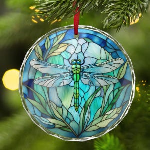 May include: A round, clear glass ornament with a dragonfly design. The dragonfly is green and blue, with a stained-glass effect. The ornament is hanging from a red ribbon, with a green background.