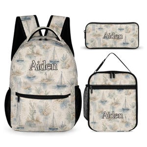 Può includere: Uno zaino, una borsa per il pranzo e un astuccio con un motivo nautico di barche a vela, fari e conchiglie nei toni del blu e del beige. Ogni articolo è personalizzato con il nome Aiden in un carattere serif marrone scuro e ha bordi neri.