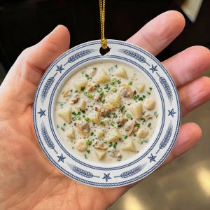 以下が含まれることがあります： ジャガイモの塊、アサリ、緑のハーブ、小さなクラッカーが入ったクリーミーなクラムチャウダーのボウルが描かれた丸いセラミック製のオーナメント。縁には青いロープ模様、麦の穂、五芒星がデザインされています。