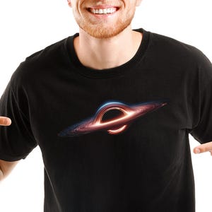Op de afbeelding: Een zwart T-shirt met een afbeelding van een zwart gat op de voorkant. Het zwarte gat wordt omringd door een ring van licht en gas.