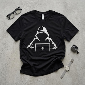 May include: Black t-shirt featuring a white graphic of a hooded figure working on a laptop with a skull and crossbones symbol. The shirt is made of a soft material and is suitable for casual wear.