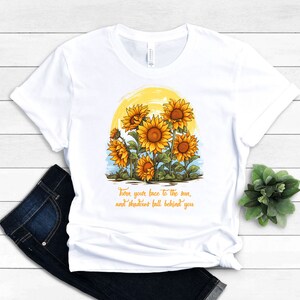 May include: White t-shirt with a graphic of sunflowers in front of a yellow sun. The text "Turn your face to the sun, and shadows fall behind you" is printed below the graphic.