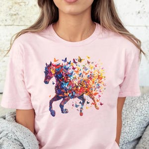 以下が含まれることがあります： 蝶々でできた馬のカラフルなグラフィックがプリントされたピンクのTシャツ。馬は走っており、蝶々は馬の周りを飛んでいます。