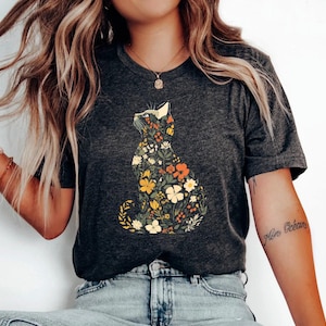 Puede incluir: Camiseta gris oscuro con un diseño floral de gato. La silueta del gato está llena de flores coloridas en tonos amarillos, naranjas y blancos. Prenda de vestir informal.