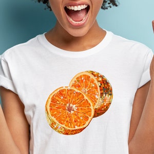 Puede incluir: Una camiseta blanca con un gráfico de dos rodajas de naranja. Una rodaja es una rodaja de naranja tradicional, mientras que la otra es una bola de discoteca.
