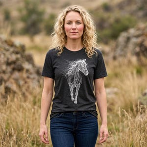 May include: Dark gray t-shirt featuring a white, stylized illustration of a horse. The horse design has flowing lines, suggesting movement. The shirt is paired with blue jeans. The overall theme is equestrian and nature-inspired.