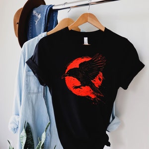 May include: Black t-shirt featuring a graphic of a black raven in front of a red circle. The raven is depicted with outstretched wings. The shirt is hanging on a wooden hanger with a blue denim shirt and a brown hat in the background.
