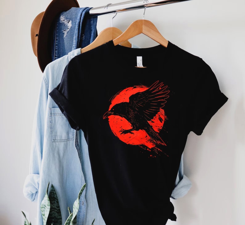 May include: Black t-shirt featuring a graphic of a black raven in front of a red circle. The raven is depicted with outstretched wings. The shirt is hanging on a wooden hanger with a blue denim shirt and a brown hat in the background.