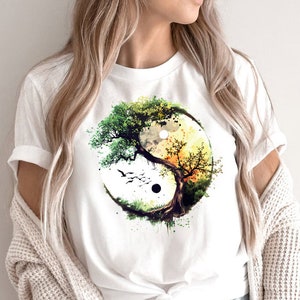 May include: White t-shirt featuring a watercolour design of a tree in the shape of a yin yang symbol. The tree has green and brown leaves and the yin yang symbol is black and white.