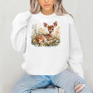 Puede incluir: Sudadera blanca con un gráfico de un cervatillo acostado en un campo de flores. El cervatillo es marrón con manchas blancas y tiene ojos grandes y marrones. Las flores son amarillas y blancas.