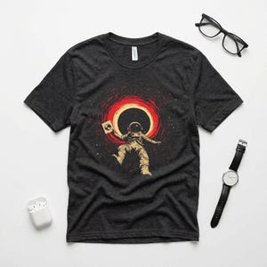 Op de afbeelding: Donkergrijs t-shirt met een astronaut die in de ruimte zweeft en een plaat vasthoudt. De astronaut draagt een ruimtepak, met een rood en geel cirkelvormig ontwerp erachter. Het t-shirt heeft een ronde hals en korte mouwen.