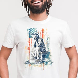 May include: A white t-shirt with a distressed graphic of the Statue of Liberty sitting with her head in her hands. The graphic is in front of an American flag with a skyscraper in the background.