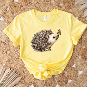May include: A yellow t-shirt with a graphic of a hedgehog and a butterfly. The hedgehog is brown and has a light brown nose. The butterfly is orange and black.