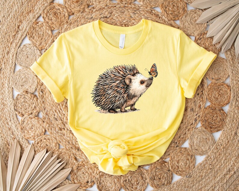May include: A yellow t-shirt with a graphic of a hedgehog and a butterfly. The hedgehog is brown and has a light brown nose. The butterfly is orange and black.