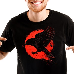 May include: Black t-shirt featuring a graphic of a black raven with outstretched wings against a red circular background. The raven has a detailed design, and the shirt is a solid color.