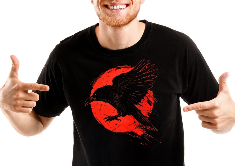 May include: Black t-shirt featuring a graphic of a black raven with outstretched wings against a red circular background. The raven has a detailed design, and the shirt is a solid color.