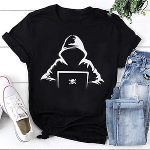Op de afbeelding: Zwart T-shirt met een witte afbeelding van een gekapte figuur die voor een laptop zit met een schedel en gekruiste beenderen op het scherm.