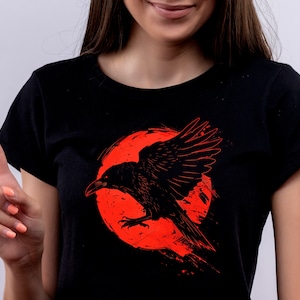 May include: Black t-shirt featuring a detailed illustration of a raven with outstretched wings against a red, circular background. The raven is depicted in a dynamic pose, with its wings spread wide. The design is printed on a black t-shirt.