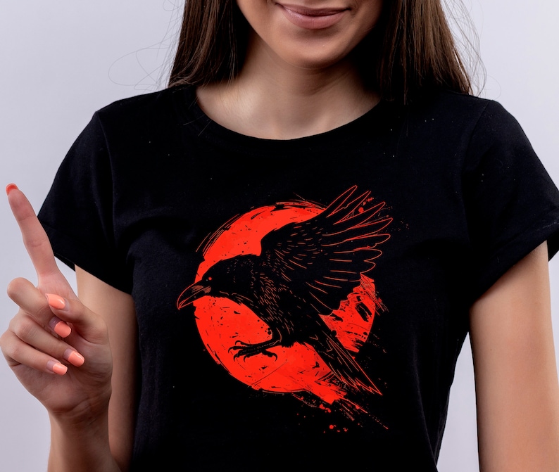 May include: Black t-shirt featuring a detailed illustration of a raven with outstretched wings against a red, circular background. The raven is depicted in a dynamic pose, with its wings spread wide. The design is printed on a black t-shirt.