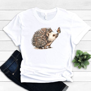May include: White t-shirt with a brown and white hedgehog looking at an orange butterfly.