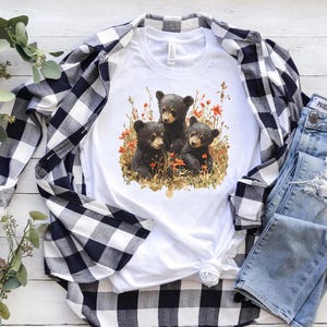 May include: White t-shirt with a black and white graphic of three bear cubs in a forest setting with red flowers. The shirt is paired with a black and white plaid shirt and blue jeans.