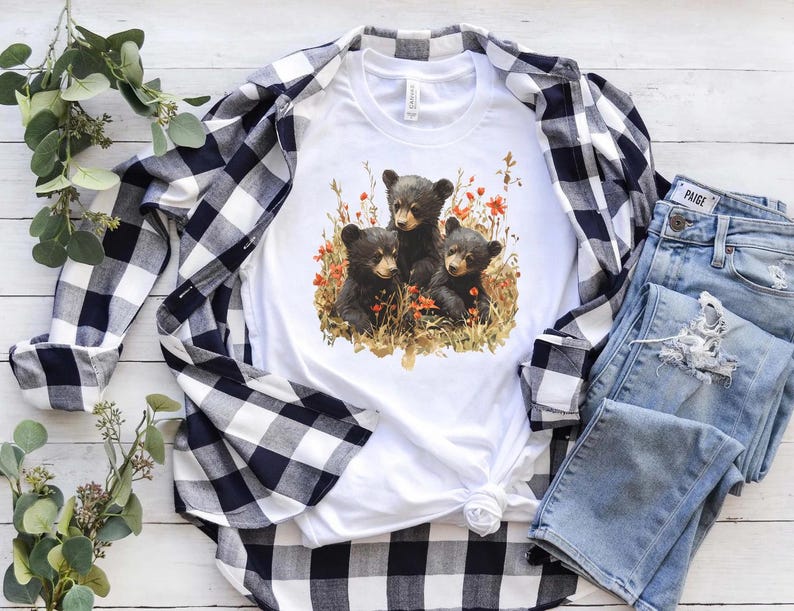 May include: White t-shirt with a black and white graphic of three bear cubs in a forest setting with red flowers. The shirt is paired with a black and white plaid shirt and blue jeans.