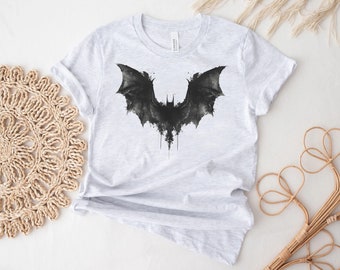 Puede incluir: Una camiseta gris con una imagen negra de estilo acuarela de una silueta de murciélago.